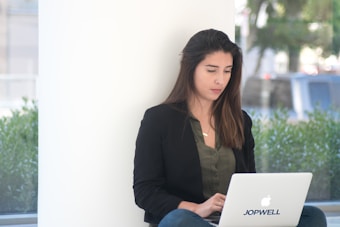 A woman sits against a white wall, using a laptop with the logo 'Jopwell' on it. She wears a dark blazer and a green shirt, with her long hair down. The environment is bright with natural light, and greenery can be seen outside the window.