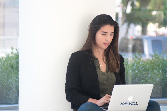 A woman sits against a white wall, using a laptop with the logo 'Jopwell' on it. She wears a dark blazer and a green shirt, with her long hair down. The environment is bright with natural light, and greenery can be seen outside the window.