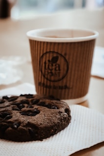 A close-up of a freshly baked chocolate chip cookie resting on a soft pink napkin with a purple measuring cup beside it