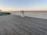 A wooden boardwalk extends along a coastal area with a metal structure resembling a sundial placed centrally. The sea is visible in the background with gentle waves, and the sky exhibits a gradient from pale orange to clear blue, suggesting a sunrise or sunset. To the left, there is a green bench and some grass, bordered by a fence.