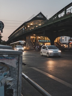 An urban evening scene featuring a street under a bridge. Several cars are moving along the road, and lights from the bridge and nearby buildings illuminate the scene. People can be seen walking up and down the stairs to the elevated train station above, which is characterized by large windows and architectural details.