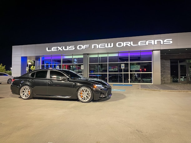 A black sedan is parked in front of a modern car dealership with the sign 'Lexus of New Orleans' illuminated above the entrance. The dealership has a stylish and sleek facade with large glass windows, and the lighting reflects a luxurious ambiance. The parking lot is well-lit at night, and the sedan has distinctive silver wheels with orange brake calipers.