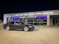 A black sedan is parked in front of a modern car dealership with the sign 'Lexus of New Orleans' illuminated above the entrance. The dealership has a stylish and sleek facade with large glass windows, and the lighting reflects a luxurious ambiance. The parking lot is well-lit at night, and the sedan has distinctive silver wheels with orange brake calipers.