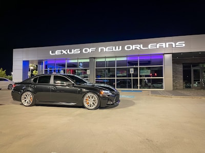 A black sedan is parked in front of a modern car dealership with the sign 'Lexus of New Orleans' illuminated above the entrance. The dealership has a stylish and sleek facade with large glass windows, and the lighting reflects a luxurious ambiance. The parking lot is well-lit at night, and the sedan has distinctive silver wheels with orange brake calipers.