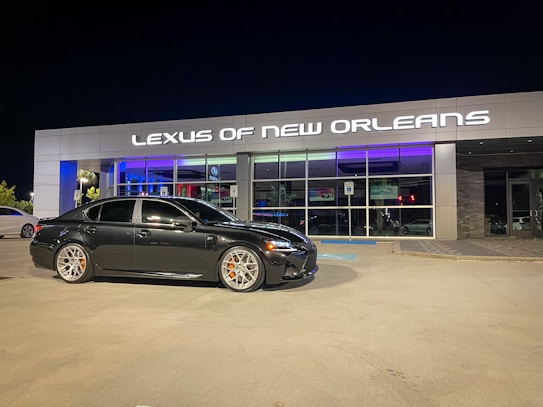 A black sedan is parked in front of a modern car dealership with the sign 'Lexus of New Orleans' illuminated above the entrance. The dealership has a stylish and sleek facade with large glass windows, and the lighting reflects a luxurious ambiance. The parking lot is well-lit at night, and the sedan has distinctive silver wheels with orange brake calipers.