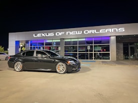 A black sedan is parked in front of a modern car dealership with the sign 'Lexus of New Orleans' illuminated above the entrance. The dealership has a stylish and sleek facade with large glass windows, and the lighting reflects a luxurious ambiance. The parking lot is well-lit at night, and the sedan has distinctive silver wheels with orange brake calipers.