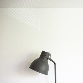 A minimalist lamp positioned against a plain, off-white wall. Soft shadows and diffused light highlight the simple and modern design of the lamp. Subtle light streaks from a window reflect on the wall surface, adding a hint of texture and depth.