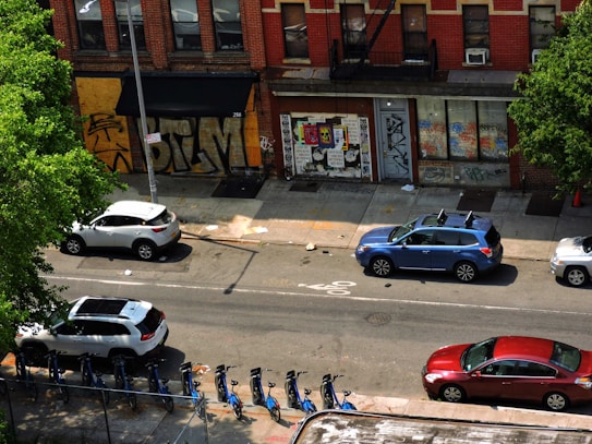 An urban street scene featuring parked cars along the side of the road with a building facade in the background. The building has graffiti-covered windows and posters. Bicycles are lined up in a bike-sharing station. Lush green trees flank the scene, providing some contrast to the urban environment.