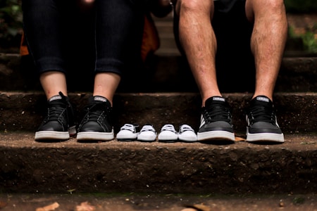 A seated couple showcasing their shoes next to a pair of small baby shoes on a stone staircase. The adults wear matching black sneakers, while the baby shoes are smaller versions in black and white.