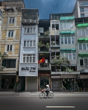 A street scene featuring a row of narrow, multi-story buildings with balconies and various signs. A Vietnamese flag is displayed on one of the buildings. A person wearing a conical hat is cycling along the street in the foreground. The buildings have a mix of colors and architectural styles, with visible air conditioning units and greenery.