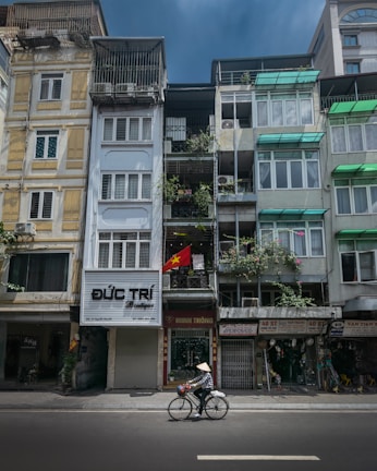 A street scene featuring a row of narrow, multi-story buildings with balconies and various signs. A Vietnamese flag is displayed on one of the buildings. A person wearing a conical hat is cycling along the street in the foreground. The buildings have a mix of colors and architectural styles, with visible air conditioning units and greenery.
