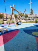 A playground area with a colorful rubberized floor, featuring a wooden swing set in the center. A child is seated on the swing. A small slide is positioned to the left. The background includes neatly trimmed bushes, trees, and a row of apartment buildings.