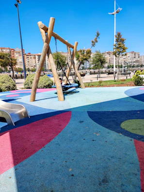 A playground area with a colorful rubberized floor, featuring a wooden swing set in the center. A child is seated on the swing. A small slide is positioned to the left. The background includes neatly trimmed bushes, trees, and a row of apartment buildings.