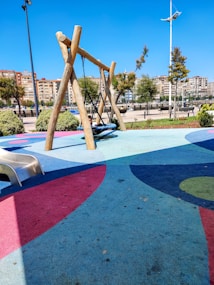 A playground area with a colorful rubberized floor, featuring a wooden swing set in the center. A child is seated on the swing. A small slide is positioned to the left. The background includes neatly trimmed bushes, trees, and a row of apartment buildings.