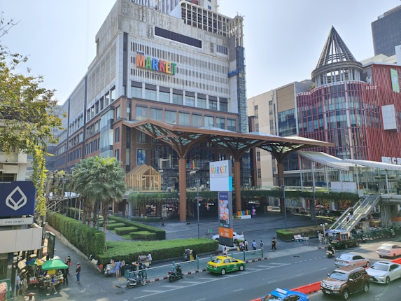 A bustling urban scene features a large, modern building with the word 'MARKET' prominently displayed on its facade. The structure is surrounded by palm trees and manicured shrubs, conveying a blend of nature and city life. In the foreground, several cars and motorcycles are visible on a busy street, alongside pedestrians and food vendors under green umbrellas. The sky is clear, allowing sunlight to highlight the architectural details.