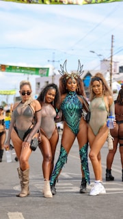 Four women dressed in elaborate and colorful costumes stand in a row on a street. They are wearing festival or carnival attire featuring sheer fabrics, sequins, and unique headpieces. The backdrop suggests an outdoor event, with banners and other festival-goers visible.