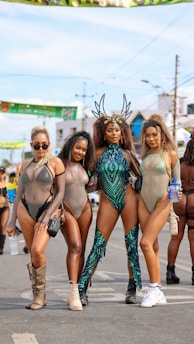 Four women dressed in elaborate and colorful costumes stand in a row on a street. They are wearing festival or carnival attire featuring sheer fabrics, sequins, and unique headpieces. The backdrop suggests an outdoor event, with banners and other festival-goers visible.