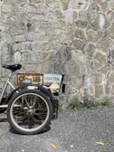A partial view of a bicycle or tricycle with a worn, rusty frame and a license plate reading 'CUSCO-PERÚ' is positioned against a textured stone wall. The vehicle's large wheel and basket are visible, surrounded by some greenery and small yellow flowers next to the rough pavement.