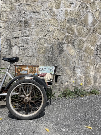 A partial view of a bicycle or tricycle with a worn, rusty frame and a license plate reading 'CUSCO-PERÚ' is positioned against a textured stone wall. The vehicle's large wheel and basket are visible, surrounded by some greenery and small yellow flowers next to the rough pavement.