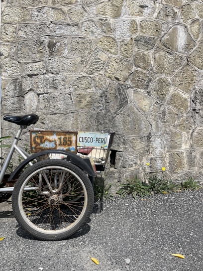 A partial view of a bicycle or tricycle with a worn, rusty frame and a license plate reading 'CUSCO-PERÚ' is positioned against a textured stone wall. The vehicle's large wheel and basket are visible, surrounded by some greenery and small yellow flowers next to the rough pavement.