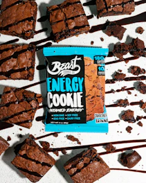 Several chocolate brownies are arranged on a light surface, drizzled with chocolate syrup. A brightly colored energy cookie package labeled Beast Cookie Co. is placed among the brownies.