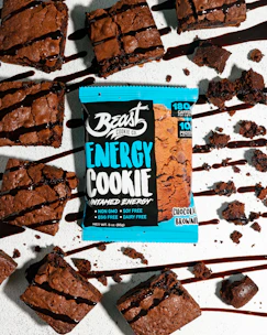 Several chocolate brownies are arranged on a light surface, drizzled with chocolate syrup. A brightly colored energy cookie package labeled Beast Cookie Co. is placed among the brownies.
