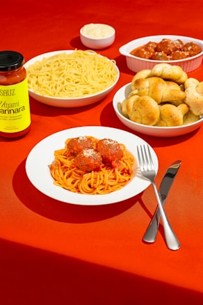 A vibrant display of Italian cuisine on a red tablecloth, featuring a plate of spaghetti and meatballs garnished with cheese. Surrounding the main dish are additional bowls, including plain pasta, garlic knots, and meatballs in sauce. A jar of marinara sauce is positioned prominently to the side, along with a fork and knife.