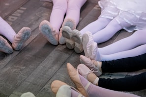 A group of people wearing ballet shoes and tights are seated in a circular arrangement on a wooden floor. The focus is on their legs and feet, which are extended outward. The tights vary in color from pink to white to black, and the shoes are mostly light pink. One individual is wearing a white tutu-like skirt.