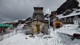 A stone temple stands amidst a snowy landscape, with colorful stripes painted on the entrance. The temple is flanked by a trident and other smaller structures. Snow blankets the surrounding area, with rock formations in the background and a cloudy sky overhead. A pathway and railing are visible to the left.