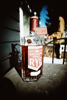 A bottle of Sagamore Spirit Rye whiskey is placed on a table with vivid lighting highlighting its amber liquid. The label on the bottle prominently displays the brand name and details about the whiskey, including that it is aged 7 years. In the blurred background, a person is visible, possibly a man is leaning casually on a railing with sunlight casting strong shadows.