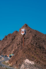 A rugged mountainous landscape with a prominent pop culture sign that says 'Hatta' twice, mounted high on a pole against a backdrop of brown rocky hills. Several vehicles, including a few colorful trucks, are visible on a winding road that curves around the base of the hills. The sky is clear and vividly blue.