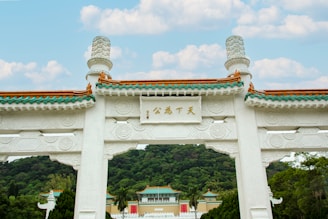 A traditional Chinese archway with intricate carvings and detailed roof tiles stands prominently in the foreground. Behind it, lush green hills and a building with similar architectural style are visible under a partly cloudy sky.