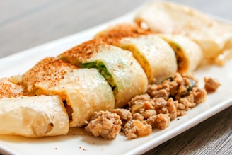 A neatly arranged dish of sliced rolls filled with vegetables and meat, garnished with a sprinkle of spices. Surrounding the rolls are cooked minced meat bits, all presented on a white rectangular plate. The background features a wooden tabletop.