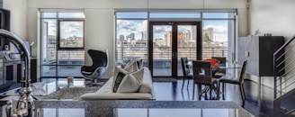 A modern living area features large windows and a glass door leading to a balcony with a cityscape view. The room is furnished with a sleek black armchair, a beige sectional sofa adorned with cushions, and a contemporary glass dining table with chairs. A staircase is visible to the side, along with a stylish cabinet. The room is spacious and filled with natural light.