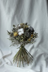 A carefully arranged bouquet of dried flowers featuring a variety of textures and colors. The bouquet contains fluffy cotton, yellow blossoms, and greenery, tied together with long green stems. It rests against a simple white fabric background, emphasizing the natural tones of the flowers.