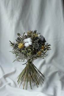A carefully arranged bouquet of dried flowers featuring a variety of textures and colors. The bouquet contains fluffy cotton, yellow blossoms, and greenery, tied together with long green stems. It rests against a simple white fabric background, emphasizing the natural tones of the flowers.