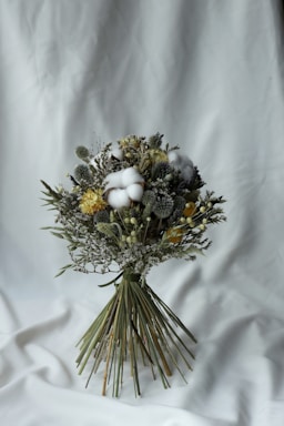 A carefully arranged bouquet of dried flowers featuring a variety of textures and colors. The bouquet contains fluffy cotton, yellow blossoms, and greenery, tied together with long green stems. It rests against a simple white fabric background, emphasizing the natural tones of the flowers.