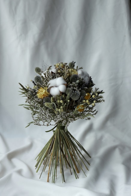 A carefully arranged bouquet of dried flowers featuring a variety of textures and colors. The bouquet contains fluffy cotton, yellow blossoms, and greenery, tied together with long green stems. It rests against a simple white fabric background, emphasizing the natural tones of the flowers.