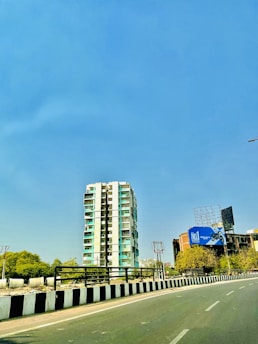 A tall residential building stands prominently against a clear blue sky. The structure is adjacent to a road lined with black and white striped barriers. To the right, a billboard advertises a product next to another building. Greenery is visible in the background, providing contrast to the urban elements.