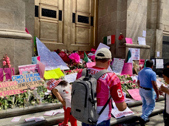 A group of people is gathered in front of an ornate doorway adorned with various protest signs and flowers. Mexican flags are visible alongside handwritten notes and messages, suggesting a public demonstration or memorial. The architecture features large stone walls and a prominent golden door. The scene is lively and filled with diverse materials on the steps.