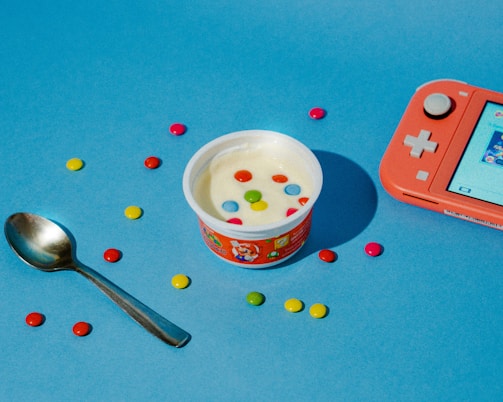 A cup of yogurt decorated with colorful candy pieces is placed on a blue surface. Surrounding the yogurt are scattered multicolored candies and a silver spoon. To the right, a coral-colored handheld gaming console is positioned, showing a display screen.