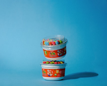 Two plastic containers are stacked, each featuring colorful candy-covered chocolates on top. The packaging has a Super Mario theme with vibrant red labels depicting Mario and other characters. The background is a solid light blue color, enhancing the playful and bright appearance of the containers.