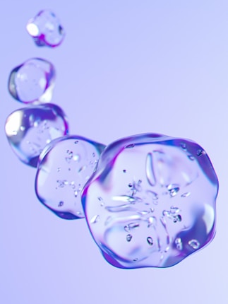 Transparent, glass-like blobs are floating in a soft gradient background of light purple and blue hues. Each blob has a smooth and rounded shape with subtle reflections and highlights.