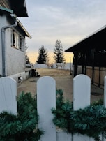 A rustic outdoor scene featuring a barn area with several horses standing near a white fence. The setting appears to be a farm or stable, surrounded by some large trees in the background. A white house is visible on the left side, with an overcast sky providing soft lighting.