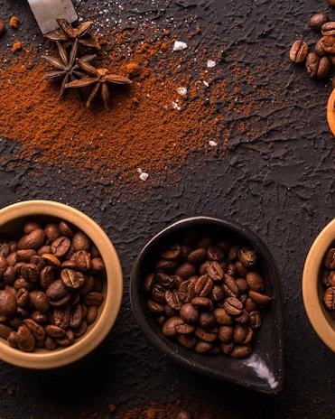 A textured dark surface is scattered with rich brown coffee grounds and whole coffee beans. Three small bowls, one black and the others tan, are filled with whole coffee beans. Star anise is placed near the powdered coffee, adding an aromatic spice element to the composition.