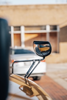 A close-up view of a side mirror attached to a vehicle, with the reflection of a yellow school bus in the mirror. A white car and a brick building are in the blurred background.