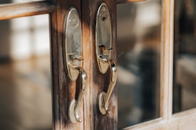 Two polished brass door handles are mounted on a wooden door with glass panels. The reflections on the metal surfaces are visible, and sunlight creates a warm glow.