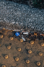 A textured surface of a sidewalk or pavement area features yellow tactile tiles with raised circular patterns. Next to this surface, a stone or gravel textured edge is visible, slightly elevating above the tactile area. Amidst the small debris, a blue plastic bottle cap and some cigarette butts are scattered on the ground.
