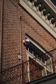 A red brick building facade featuring a distinctive white cornice at the top. A surveillance camera is mounted above a large wooden door, which is flanked by a vintage-style exterior light fixture. An ornate railing runs horizontally across the lower part of the image.