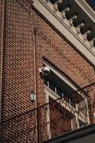 A red brick building facade featuring a distinctive white cornice at the top. A surveillance camera is mounted above a large wooden door, which is flanked by a vintage-style exterior light fixture. An ornate railing runs horizontally across the lower part of the image.